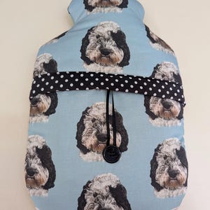 Puede incluir: Una funda para bolsa de agua caliente de color azul claro con un patrón repetitivo de caras de perro en blanco y negro. La funda incluye una banda horizontal de tela negra con lunares blancos, asegurada por un cordón negro y un botón negro.