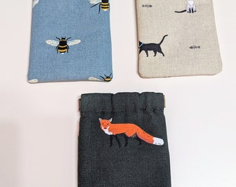 Pode incluir: Três pequenas bolsas de cordão com padrões diferentes. A primeira bolsa é azul com um padrão repetido de abelhas amarelas e pretas. A segunda bolsa é bege com um padrão repetido de gatos pretos e espinhas de peixe. A terceira bolsa é verde escura com um padrão repetido de raposas laranja.