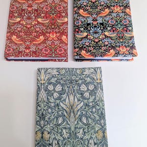 May include: Three rectangular notebooks featuring intricate botanical and bird patterns. One notebook has a red background, one has a black background, and the third features a blue-grey floral design with symmetrical leaf and petal motifs.