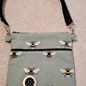 Può includere: Una borsa a tracolla blu chiaro con una tracolla nera e una chiusura a zip. La borsa è decorata con un motivo ripetitivo di api gialle e nere. La borsa ha un anello argentato con un inserto in tessuto a pois neri e bianchi.