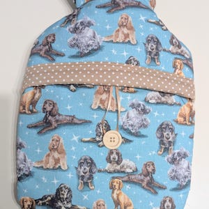 Puede incluir: Una funda para botella de agua caliente azul con un ribete de lunares marrones y blancos. La funda presenta un patrón repetido de perros sobre un fondo azul con estrellas blancas. Un botón de madera está unido a la funda.
