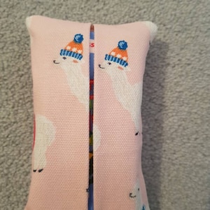 Pode incluir: Uma capa de caixa de len&ccedil;os de tecido rosa com um design de duas lhamas brancas usando chap&eacute;us de inverno azuis e laranja.