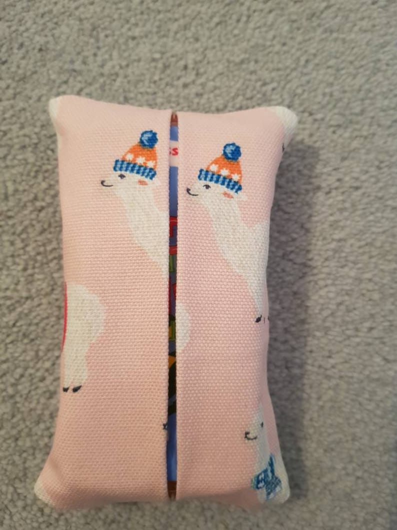 Pode incluir: Uma capa de caixa de len&ccedil;os de tecido rosa com um design de duas lhamas brancas usando chap&eacute;us de inverno azuis e laranja.