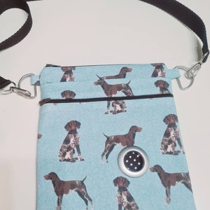 Può includere: Una borsa a tracolla blu chiaro con una tracolla regolabile marrone e una chiusura a zip. La borsa è decorata con un motivo ripetitivo di Bracchi tedeschi a pelo corto marroni e bianchi. La borsa ha una piccola ventilazione rotonda argentata con pois neri.