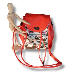 Può includere: Uno zaino rosso e bianco con motivo, con una patta rossa e una chiusura a zip. Lo zaino ha spallacci regolabili e un manico superiore.