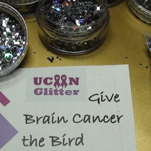 May include: A close-up of four containers of silver glitter with iridescent accents. The containers are labeled "U Can Glitter" and "Give Brain Cancer the Bird".