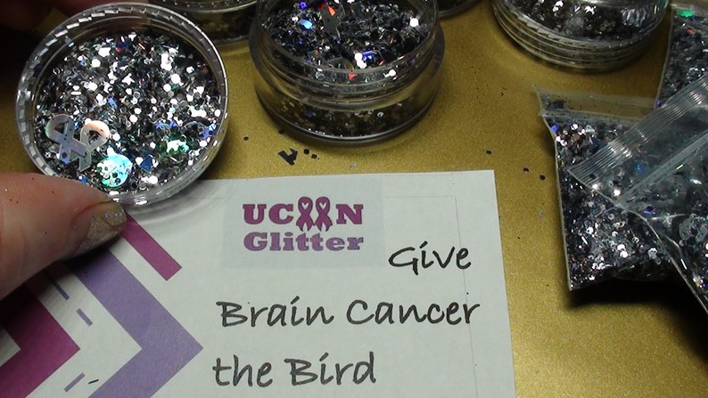 May include: A close-up of four containers of silver glitter with iridescent accents. The containers are labeled "U Can Glitter" and "Give Brain Cancer the Bird".
