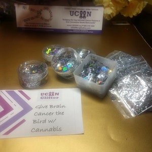 May include: A small card with the text "Give Brain Cancer the Bird w/ Cannabis" and the words "UCAN Glitter" above it. There are five small containers of silver glitter, some in clear jars and some in plastic containers.