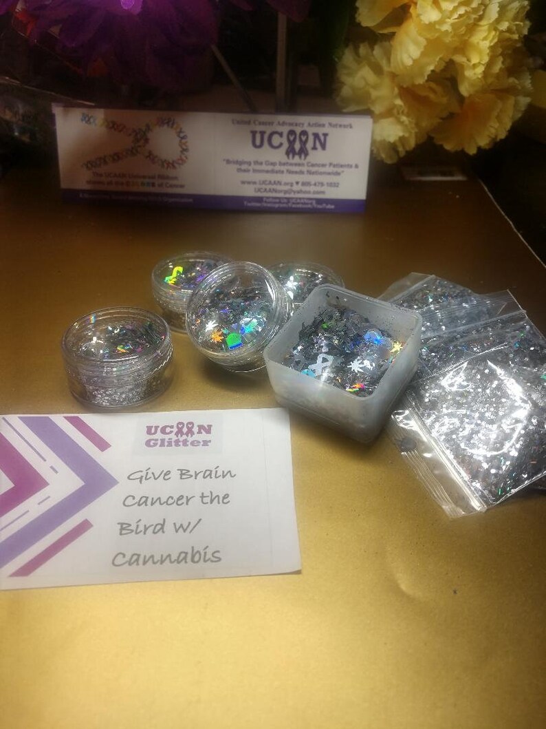 May include: A small card with the text "Give Brain Cancer the Bird w/ Cannabis" and the words "UCAN Glitter" above it. There are five small containers of silver glitter, some in clear jars and some in plastic containers.