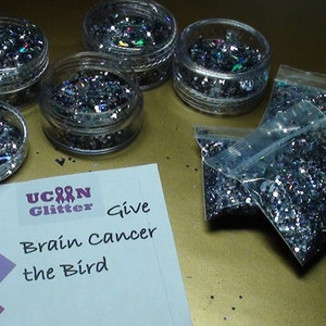 May include: A collection of silver glitter in small jars and plastic bags. The jars and bags are on a gold surface. A white card with the text "U Can Glitter Give Brain Cancer the Bird" is in the lower left corner of the image.