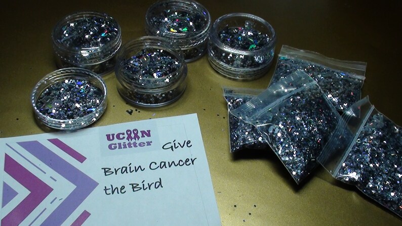 May include: A collection of silver glitter in small jars and plastic bags. The jars and bags are on a gold surface. A white card with the text "U Can Glitter Give Brain Cancer the Bird" is in the lower left corner of the image.