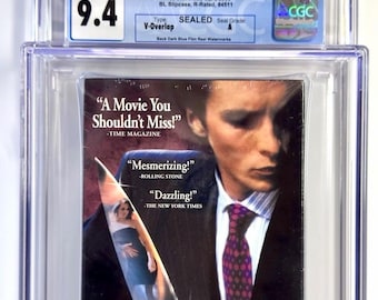 Puede incluir: Una copia VHS sellada de la película "American Psycho", calificada con 9.4 por CGC. La portada presenta a un hombre sosteniendo un cuchillo con una mujer reflejada en la hoja. Incluye citas de Time Magazine, Rolling Stone y The New York Times.
