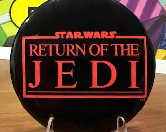 Puede incluir: Un botón redondo negro de Star Wars con el texto "STAR WARS RETURN OF THE JEDI" en rojo. El botón se muestra en un soporte de plástico transparente, con un fondo colorido de regalos envueltos.