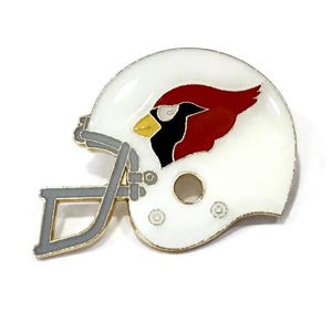 May include: A white football helmet pin with a red cardinal bird logo. The helmet has gray face mask and trim, and a gold-colored rim. The cardinal bird has a black head, yellow beak, and red body.