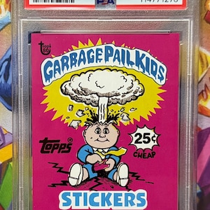 K&ouml;nnte beinhalten: Sammelkarte der Garbage Pail Kids von 2018, mit einer Cartoonfigur und einer Pilzwolkenexplosion. Die Karte befindet sich in einer Schutzh&uuml;lle und enth&auml;lt den Text "STICKERS".