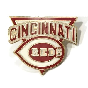 May include: A Cincinnati Reds pin with a red, white, and gold color scheme. The pin features the team name in a stylized font, with the word "REDS" in a white oval. The pin is a triangular shape with a red border.