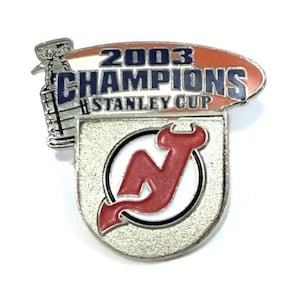 May include: A silver-toned pin featuring the text "2003 CHAMPIONS STANLEY CUP" in blue and white, with a red and white oval background. The pin also displays a red "N" logo within a circle and a silver Stanley Cup trophy.