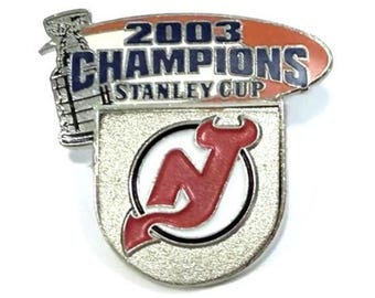 Może przedstawiać: Srebrna przypinka z napisem "2003 CHAMPIONS STANLEY CUP" w kolorze niebiesko-białym, na czerwono-białym owalu. Przypinka przedstawia również czerwone logo "N" w okręgu i srebrne trofeum Pucharu Stanleya.