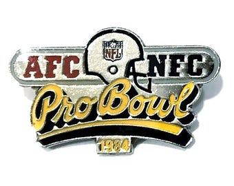Puede incluir: Un pin plateado del Pro Bowl con el texto "AFC" en rojo y "NFC" en negro. El pin presenta un casco de fútbol americano y el texto "Pro Bowl" en amarillo con un contorno negro. El año "1984" está en la parte inferior.