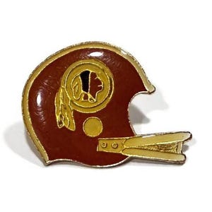 May include: A vintage football helmet pin with a maroon and gold color scheme. The helmet features a gold emblem of a Native American profile. The pin has a gold border and a gold facemask.