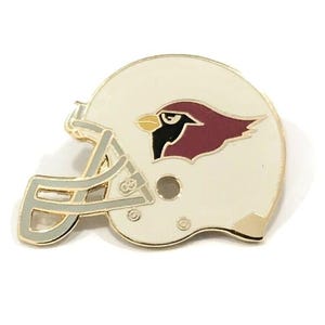 May include: A cream-colored football helmet pin with a gold-colored trim. The helmet features the Arizona Cardinals logo, a cardinal bird in maroon and black. The pin is a sports-themed accessory.