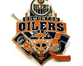 Puede incluir: Un pin con el logotipo de los Edmonton Oilers. El pin tiene forma de escudo con el nombre y el logotipo del equipo en naranja, azul y blanco. Incluye un palo de hockey, una máscara de portero y el logotipo de la NHL.