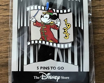 Puede incluir: Un pin en blanco y negro con Mickey Mouse vestido de director de orquesta. El pin tiene forma de tira de película con el año 1935 en él. El pin es parte de una serie llamada "Countdown to the Millennium 2000". El pin es de The Disney Store.