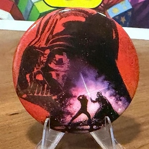 May include: A round, red button featuring Darth Vader's silhouette and a scene from Star Wars. The button is displayed on a clear plastic stand, with a colorful background visible behind it.