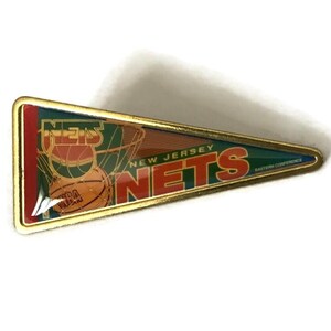 May include: A gold-colored pin shaped like a pennant with the words "New Jersey Nets" and a basketball in a hoop. The pin has a red, orange, green, and blue color scheme.