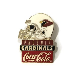 May include: A gold-colored pin featuring a white football helmet with a red cardinal bird logo. The helmet sits on top of a red, white, and black sign that reads "Phoenix Cardinals Coca-Cola".