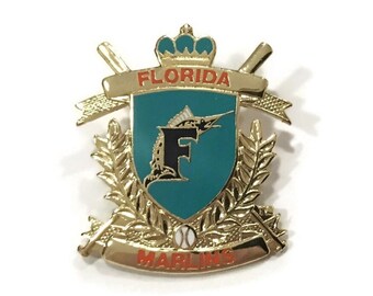Puede incluir: Pin de color dorado con un escudo azul que presenta una "F" negra y un pez marlin. El escudo está rodeado por una corona de laurel dorada y una pancarta que dice "FLORIDA MARLINS".