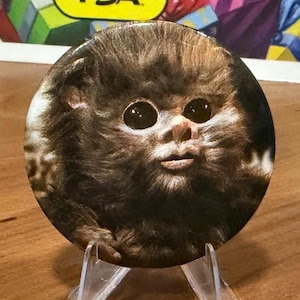May include: A round button featuring a furry creature with large, dark eyes and a small mouth. The button is brown and black, and is displayed on a clear plastic stand. The background includes colorful gift boxes and the text "PSA".