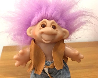 Puede incluir: Una muñeca troll vintage con una cara sonriente, orejas grandes y un mechón de pelo morado. La muñeca lleva un chaleco marrón y pantalones cortos de mezclilla. La muñeca está sobre una superficie de madera.