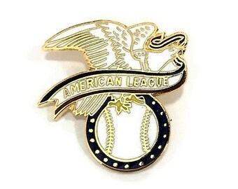 Puede incluir: Un pin dorado con un águila blanca con las alas extendidas, una pancarta con la inscripción "AMERICAN LEAGUE" y una pelota de béisbol. La pelota es blanca con costuras doradas y un borde azul marino.