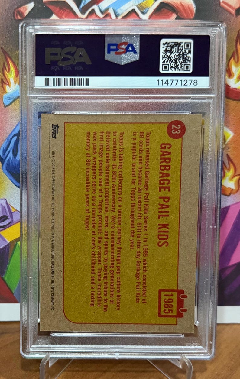 K&ouml;nnte beinhalten: Eine bewertete Garbage Pail Kids Sammelkarte von 1985 mit dem Serientitel und der Nummer 23. Die Karte befindet sich in einer Schutzh&uuml;lle mit einem PSA-Authentifizierungsetikett.