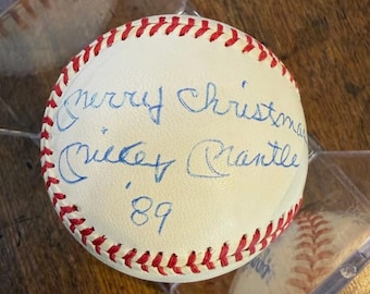 Puede incluir: Una pelota de béisbol blanca con costuras rojas y texto manuscrito azul que dice "Merry Christmas Mickey Mantle '89". La pelota está en una funda protectora transparente, con otra pelota visible en el fondo.