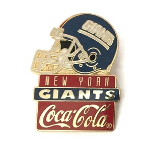 May include: A New York Giants and Coca-Cola pin. The pin features a blue football helmet with the word "GIANTS" and the team logo. Below the helmet is a red rectangle with "NEW YORK GIANTS" and "Coca-Cola" in white lettering.