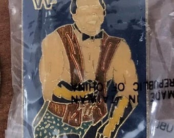 Puede incluir: Un pin rectangular con un luchador con chaleco y pajarita, con el texto "BRUTUS BEEFCAKE" en la parte inferior. El pin tiene un fondo azul con las letras "WF" en la parte superior. El pin está en una bolsa de plástico transparente.