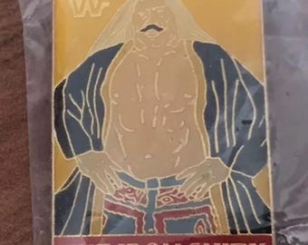 Puede incluir: Un pin rectangular con una imagen estilizada de un hombre con bigote y vestimenta tradicional de Oriente Medio. El pin tiene un fondo dorado con el texto "THE IRON SHIEK" en rojo en la parte inferior y el logotipo de la WWF en la parte superior.