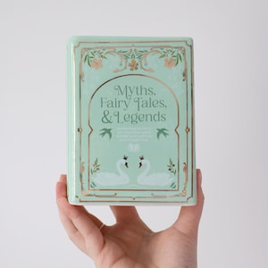 May include: A mint green book titled "Myths, Fairy Tales, & Legends" with gold accents and floral designs. The cover features two white swans and decorative elements. The book is held in a hand.