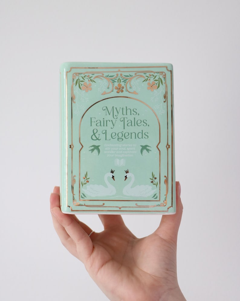 May include: A mint green book titled "Myths, Fairy Tales, & Legends" with gold accents and floral designs. The cover features two white swans and decorative elements. The book is held in a hand.
