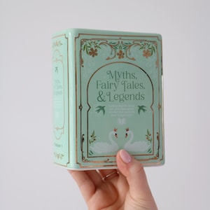 May include: A mint green book-shaped box with gold accents and the title "Myths, Fairy Tales, & Legends." The box features illustrations of swans and floral designs. The box is held in a person's hand.