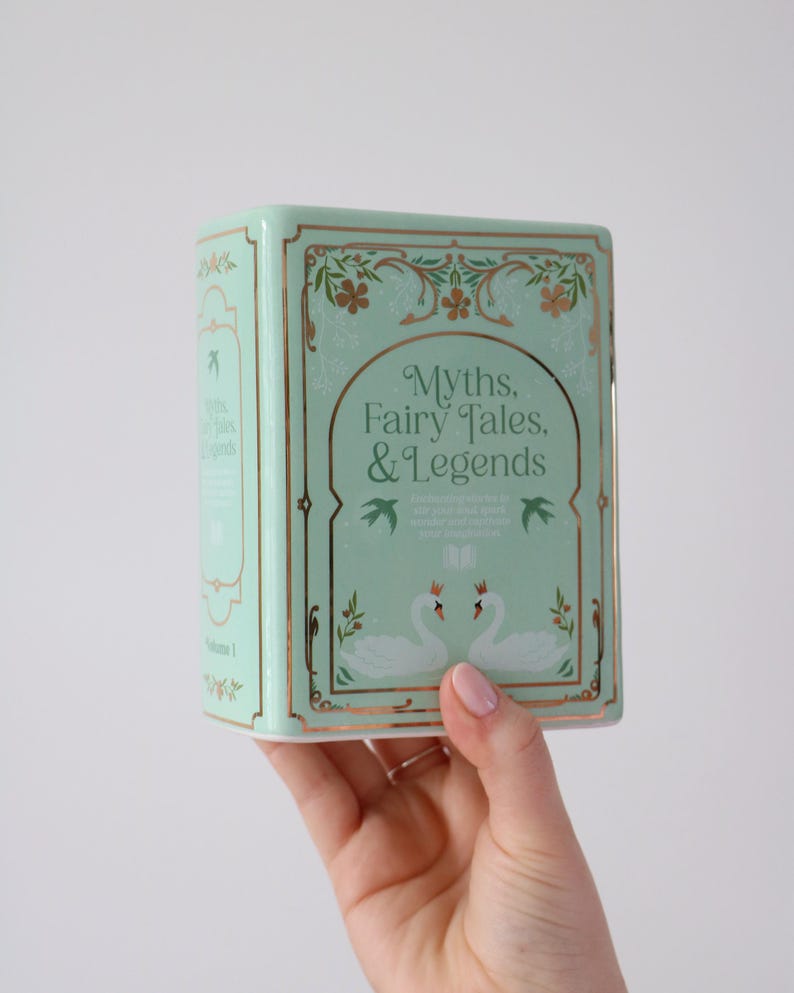 May include: A mint green book-shaped box with gold accents and the title "Myths, Fairy Tales, & Legends." The box features illustrations of swans and floral designs. The box is held in a person's hand.