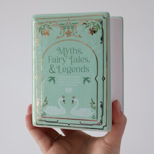 May include: A mint green ceramic book with gold accents, featuring the title "Myths, Fairy Tales, & Legends." The cover has a decorative border, swan illustrations, and the text "Enchanting stories to stir your soul, spark wonder and captivate your imagination."