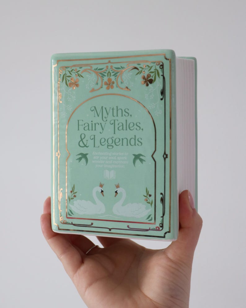 May include: A mint green ceramic book with gold accents, featuring the title "Myths, Fairy Tales, & Legends." The cover has a decorative border, swan illustrations, and the text "Enchanting stories to stir your soul, spark wonder and captivate your imagination."