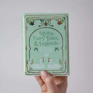 May include: A rectangular, mint-green book titled "Myths, Fairy Tales, & Legends." The cover features gold accents, floral designs, and two swans. The book's design is reminiscent of a vintage aesthetic.