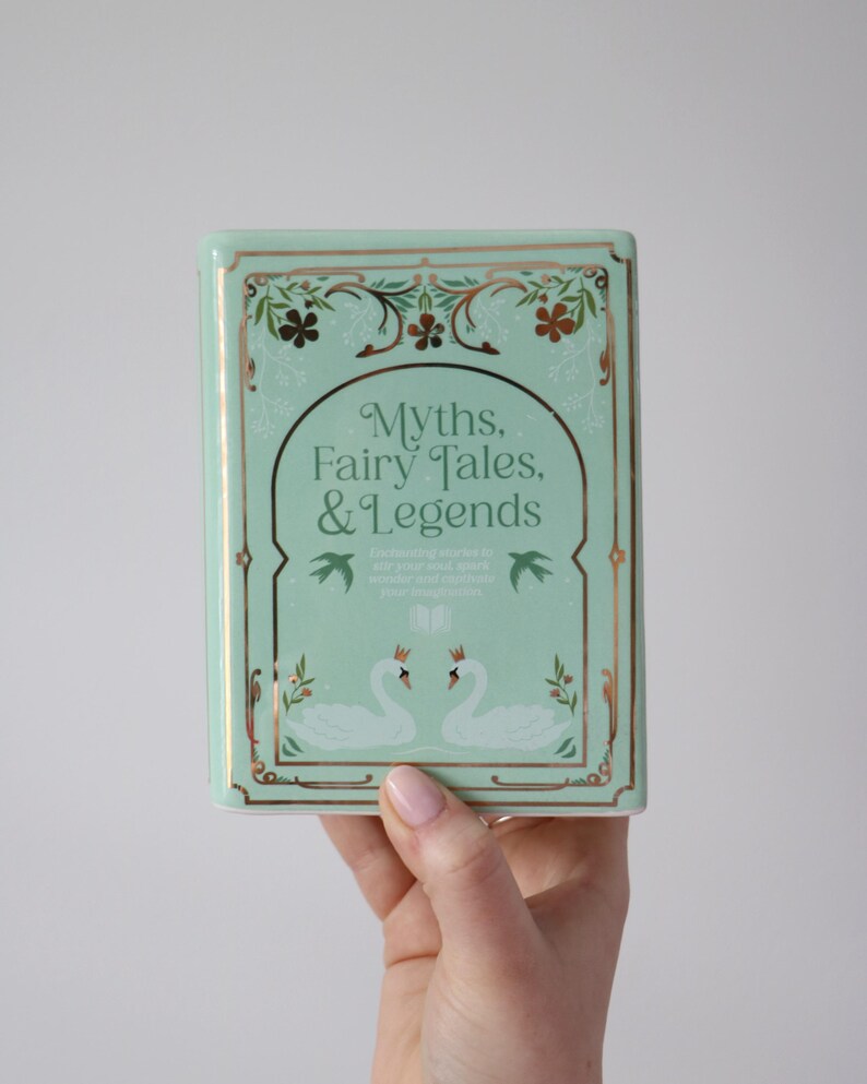 May include: A rectangular, mint-green book titled "Myths, Fairy Tales, & Legends." The cover features gold accents, floral designs, and two swans. The book's design is reminiscent of a vintage aesthetic.