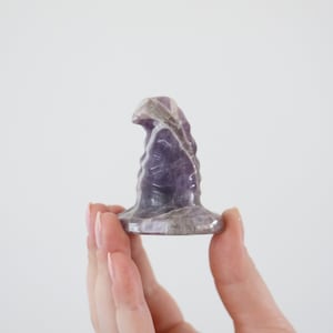 May include: A carved amethyst crystal sculpture with a rounded base and a textured, vertical form. The sculpture is held in a hand against a white background. The crystal features shades of purple and white.