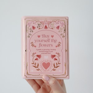 May include: A pink book with gold edging and the text "Buy yourself the flowers." The book features a floral design and a heart. The subtitle reads "A guide to growing, healing and learning to put yourself first."