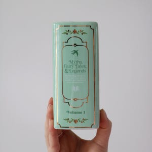 May include: A rectangular, mint-green ceramic container with gold trim and the text "Myths, Fairy Tales, & Legends, Volume 1." The design includes floral and bird motifs, suggesting a literary theme.